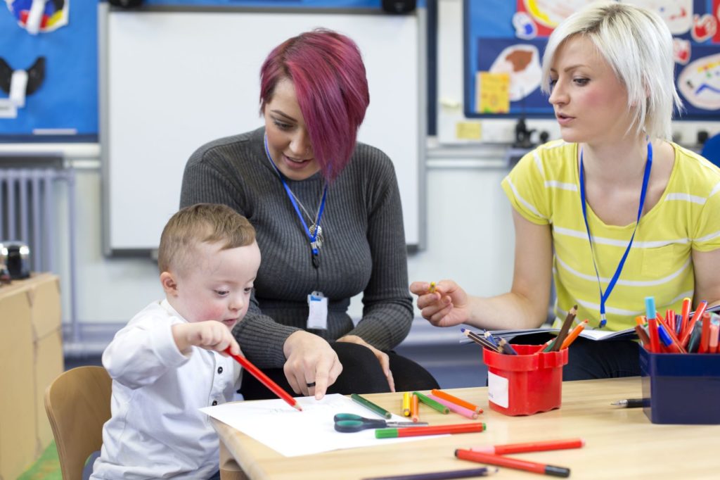 an early childhood educator and early childhood education assistant help a child draw with a red pencil