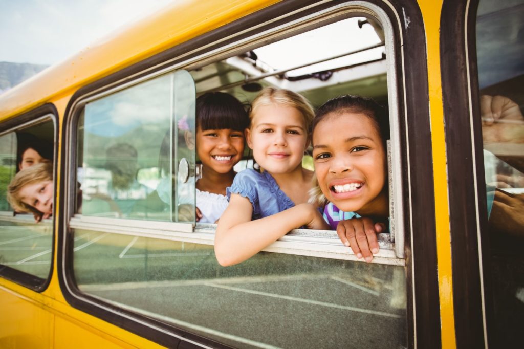 three young children smile while looking out of a school bus window