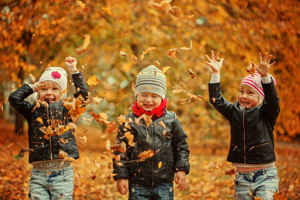 three children wearing leather jackets and beanies throw handfuls of fall leaves into the air