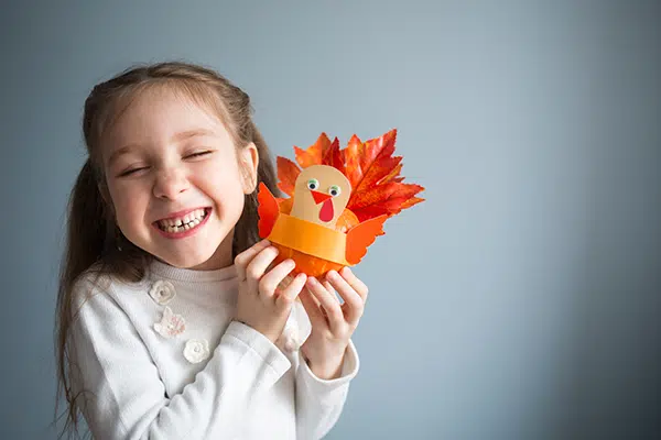 a little girl smiles while holding up a turkey she made out of paper and leaves